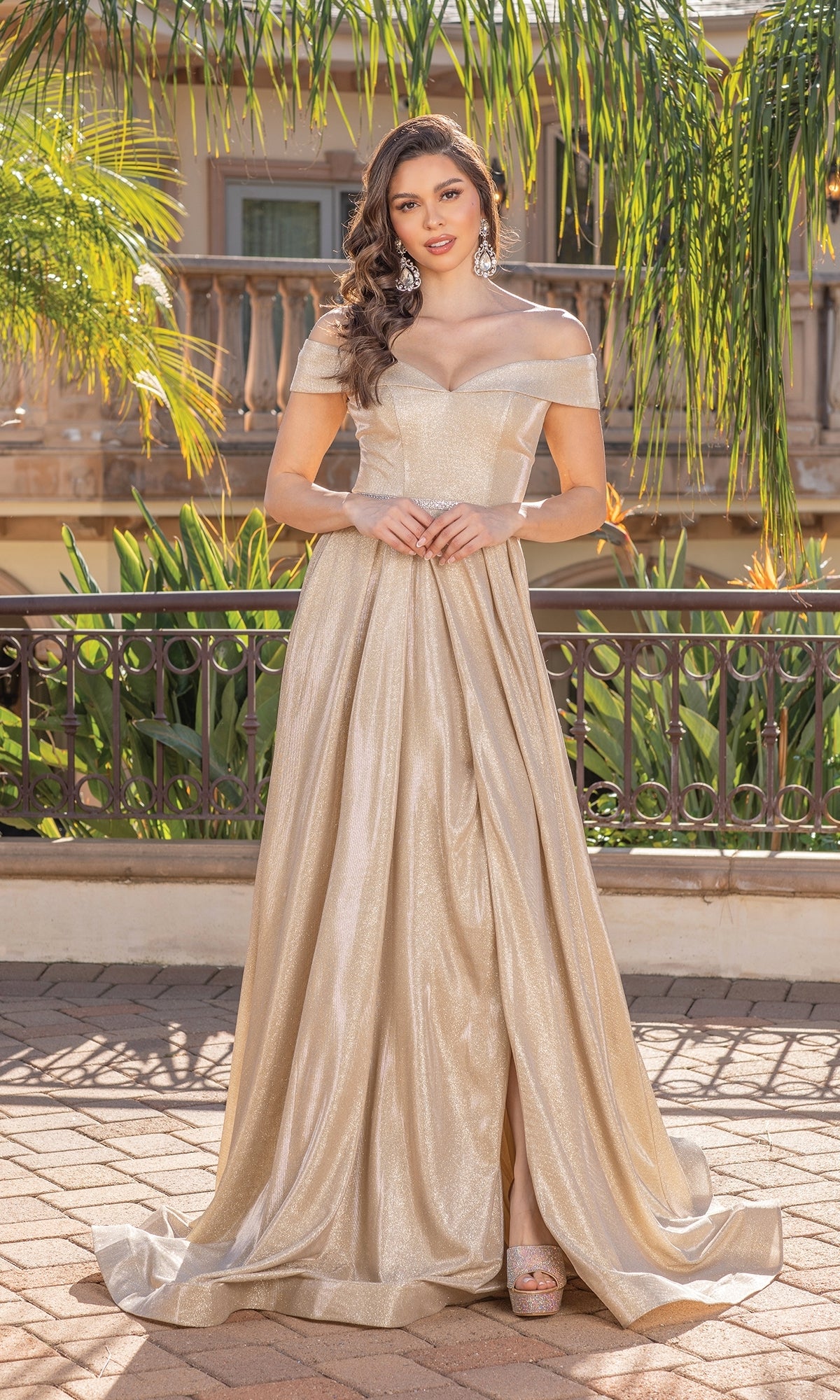 A woman poses outdoors in the Off-Shoulder Long Prom Dress: Dancing Queen 2824—an elegant glitter gown with a full skirt, accessorized with statement earrings and platform heels, surrounded by greenery and a building.